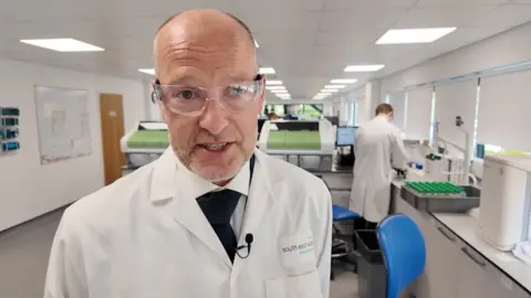 A man in safety glasses and a white coat stands in a laboratory with other scientists working in the background