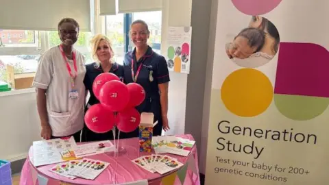South Tyneside and Sunderland NHS Foundation Trust (STSFT) Research Assistant Practitoner Folasade Olufemi-Ajayi and Research Midwives Lucy Rowland and Kirsten Herdman smiling in front of a table with leaflets about the study. There is colourful banner reading Generation Study next to them. Folasade Olufemi-Ajayi has short black hair and glasses. Lucy Rowland's blonde hair is in bun. Kirsten Herdman's brown hair is also in a bun at the back of her head. They are all wearing medical scrubs.
