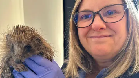 Andy Longhurst Sharon is holding up a hedgehog to the camera. She has shoulder-legth blonde hair and is wearing black rimmed glasses. She is smiling at the camera and wearing blue rubber gloves.
