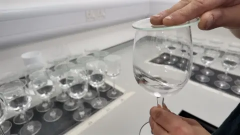 BBC A wine glass containing water is held in one hand while a small glass disc is placed over it to gather the odour of the sample