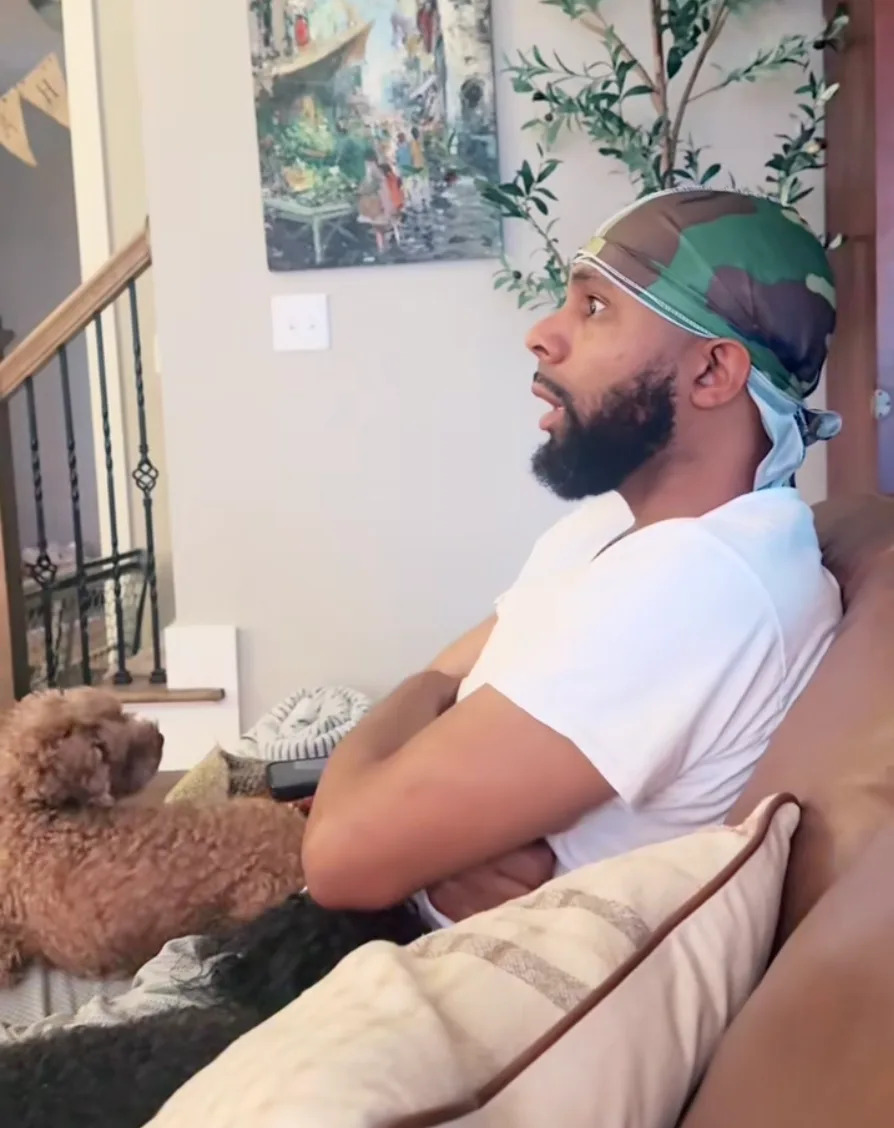 A man sits on a couch, wearing a durag and a T-shirt, with a dog beside him