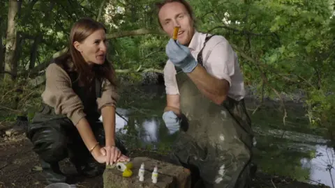 A woman on the left with long dark brown straight hair, sat at a rock with small bottles and a yellow ammonia testing kit balanced on it. On the right is a man, with blond hair, tied back, showing a test tube of water. He wears a white shirt and a khaki apron. In the background is a river, with tree overhanging the water.
