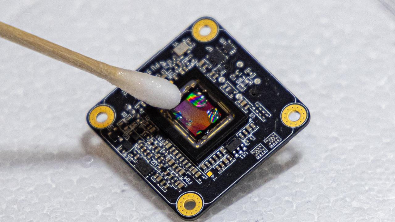 A black square with printed circuitry, tilted at an angle, against a white background. A wet q-tip points at the center of the square.