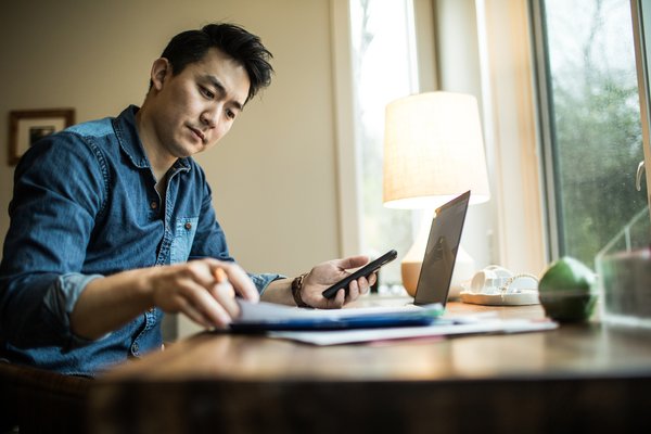 Person sits at window while working on laptop, paperwork and phone.
