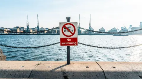 Getty Images A “No swimming” sign fixed to railings in front of a dock in east London, with water, industrial cranes, and rows of warehouses and flats visible in the background.