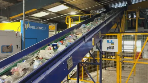 Plastic containers are travelling up a conveyor belt inside a large warehouse.