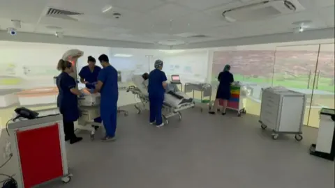 Hospital staff are working in the simulation room. An interactive screen around the room creates the environment they are practising for. There is a bed and other equipment in the room itself.