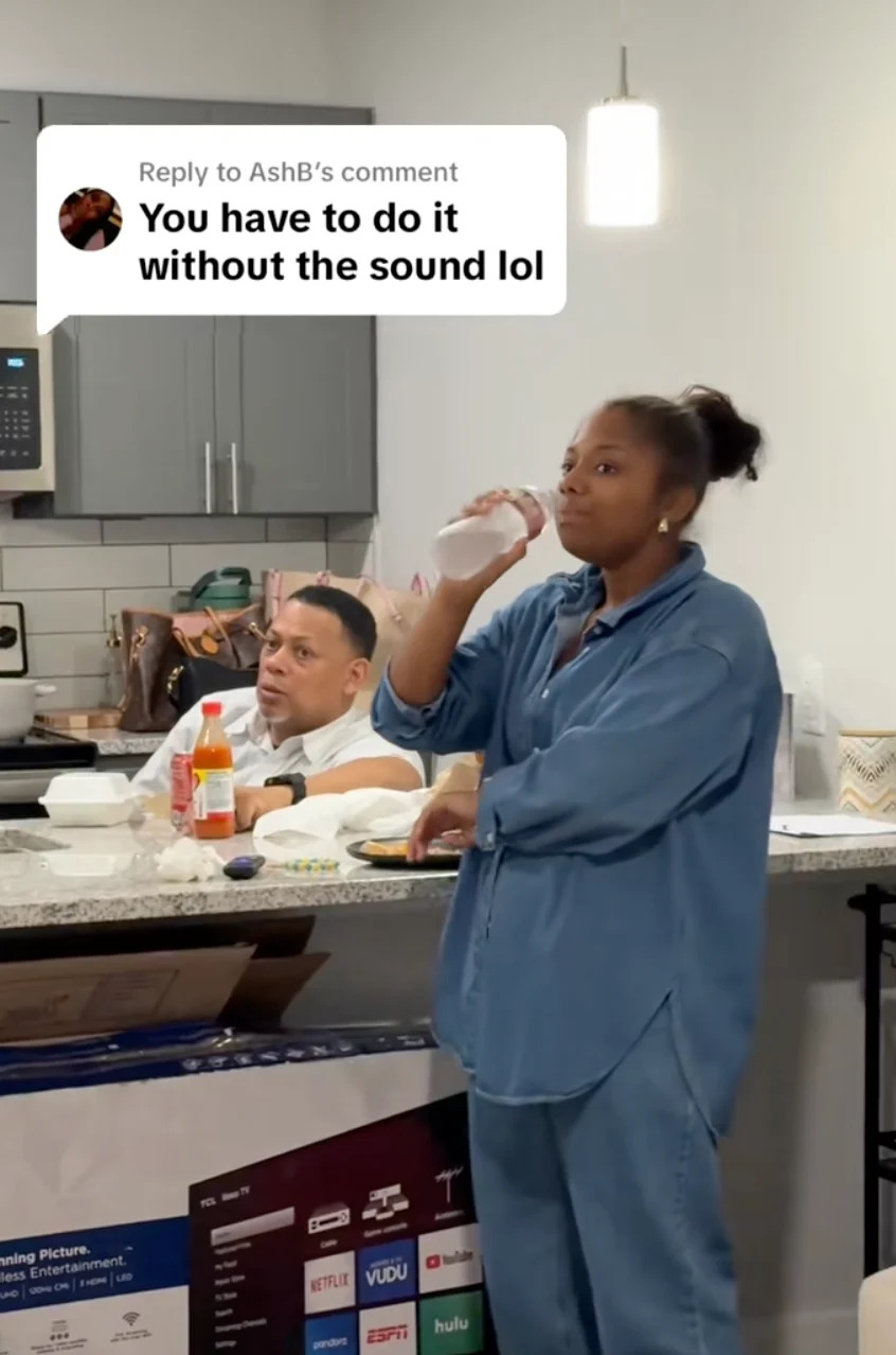 A woman drinks from a bottle in a kitchen with a man sitting behind her. A comment overlay reads, "You have to do it without the sound lol."