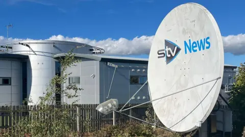 BBC STV News headquarters featuring a large satellite dish, modern architecture, and a clear blue sky with scattered clouds.