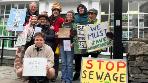 Jonah Fisher/BBC A group of anti-sewage campaigners pose for a photo. They are holding signs saying things such as "Stop Sewage" and "We must Save Windermere".