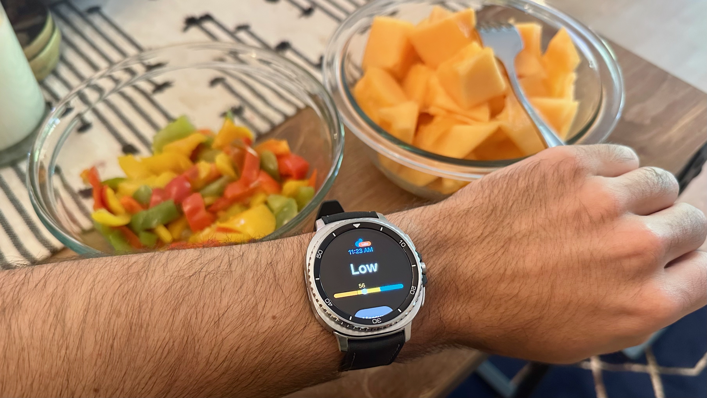 The Samsung Galaxy Watch 8 Classic on a person's wrist showing the Antioxidant Index widget with a "Low" score, held in front of two bowls containing colorful fruits and veggies.