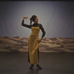A woman wearing a mustard-colored dress holds one of her hands to her head. Behind her is a backdrop with an image of an arid landscape.