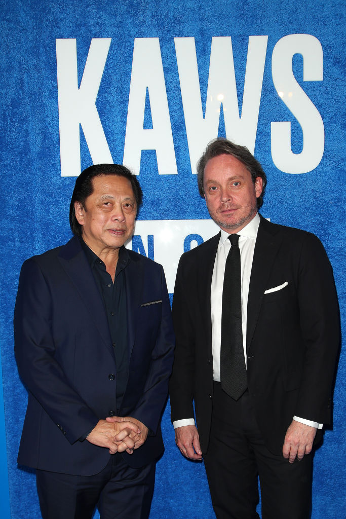 Two men in suits beneath a sign that says 'KAWS.'