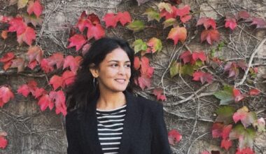 A woman poses for a photo in front of a wall covered in vines and red leaves. She