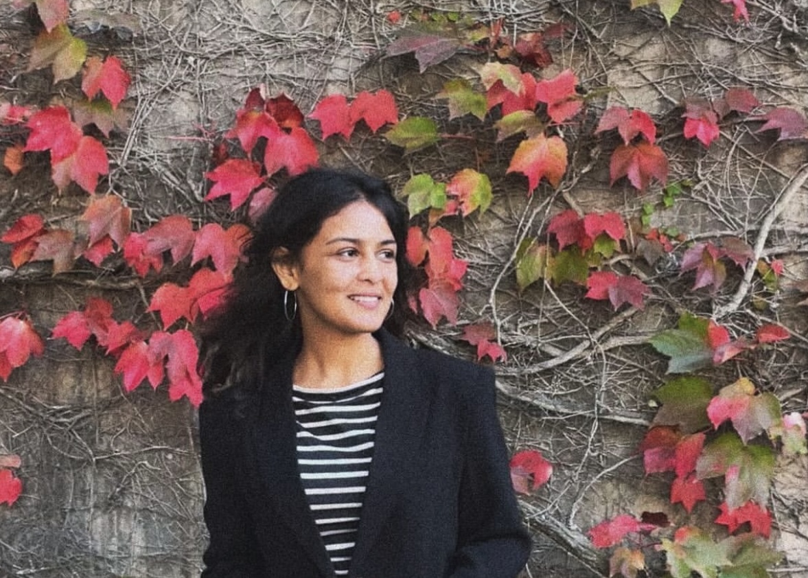 A woman poses for a photo in front of a wall covered in vines and red leaves. She
