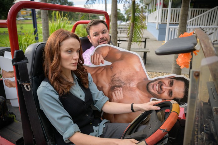 Anna Samson and Lloyd Griffith in Return to Paradise. They are sat in a car, Samson is driving and Griffith is holding a life size cardboard cutout of a topless Glenn, the character played by Tai Hara.