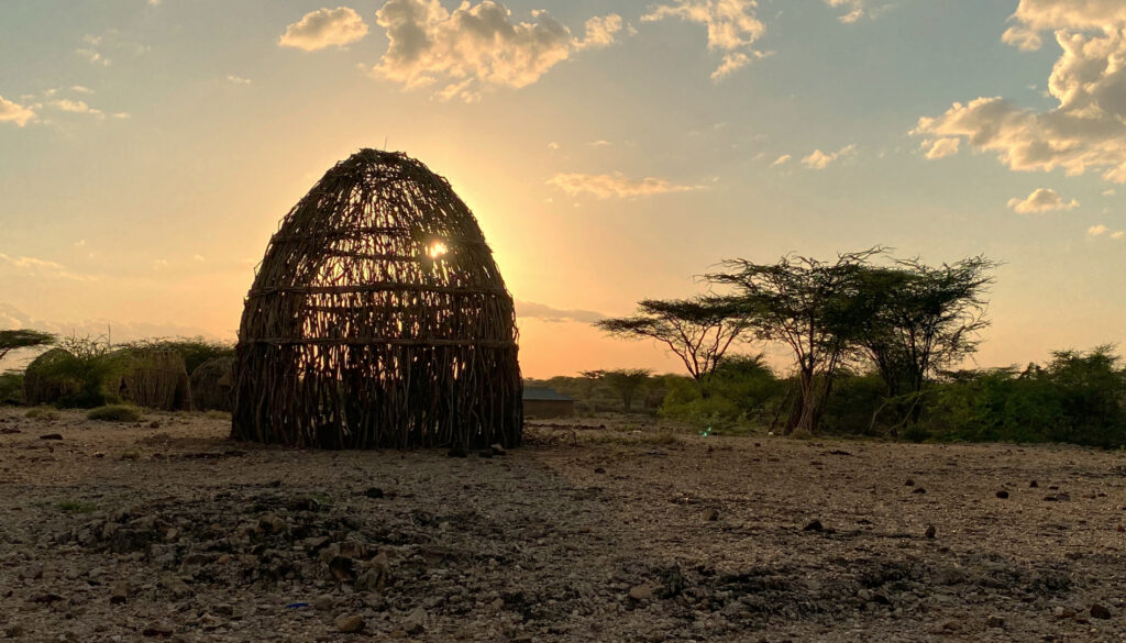a setting sun shines through a conical hut made of branches, a light blue sky in the distance