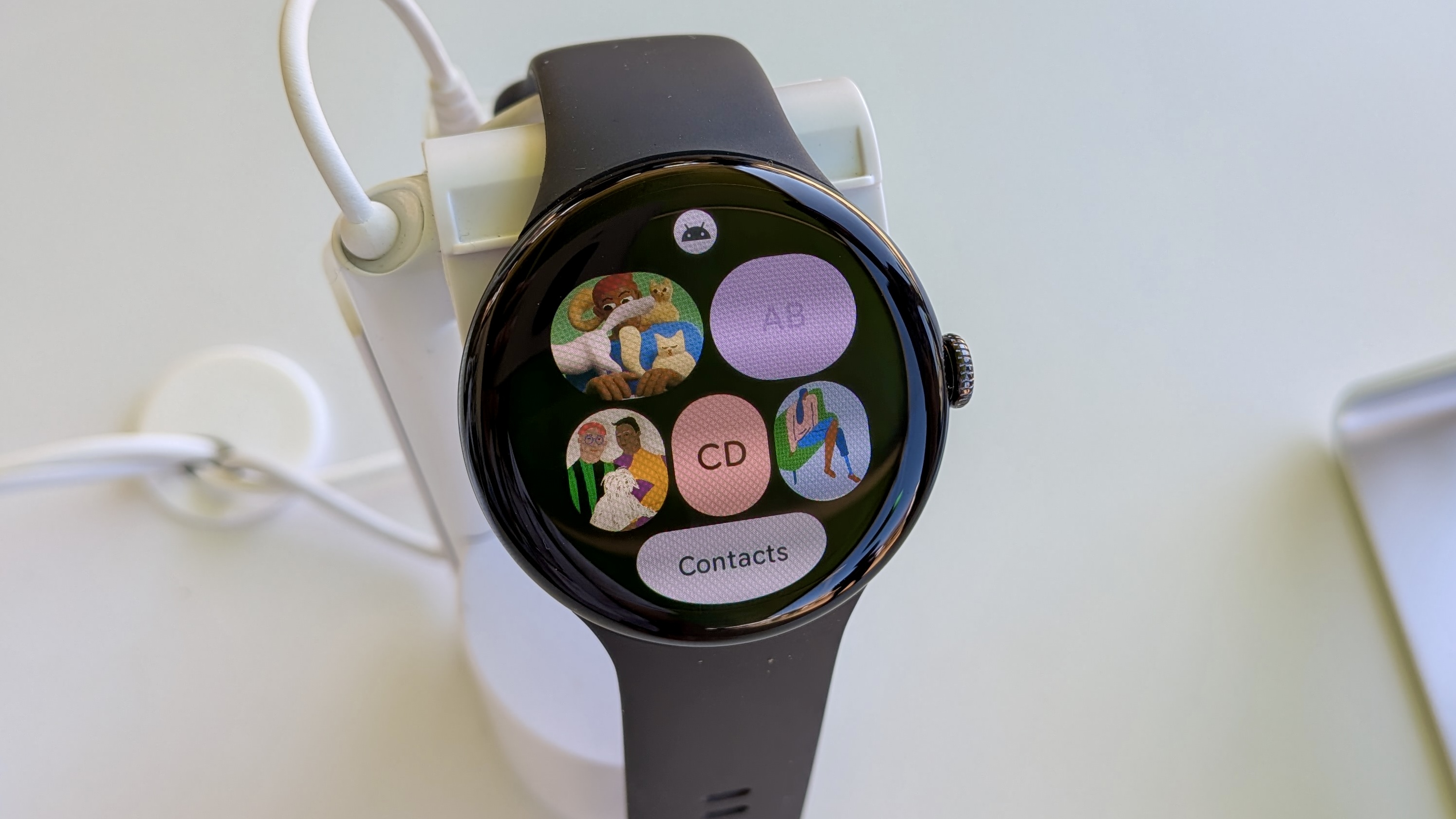 The Contacts Tile on Wear OS 6 on the Pixel Watch 3, showing different contact icons for individual numbers and an All Contacts button on the bottom edge.