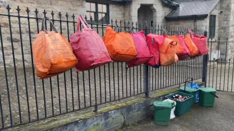 BBC Five pairs of orange and red recycling bags are hanging from wrought iron railings. The property behind the railings is made of stone and is reminiscent of an old school or chapel. Below the bags are three green food caddies, one box full of beer bottles and a blue recycling bag for paper.
