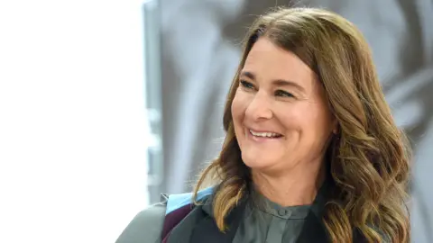 Jamie McCarthy/Getty Images Melinda French Gates with wavy brown hair smiling warmly, wearing a dark green blouse under a black vest, sitting indoors with bright light in the background