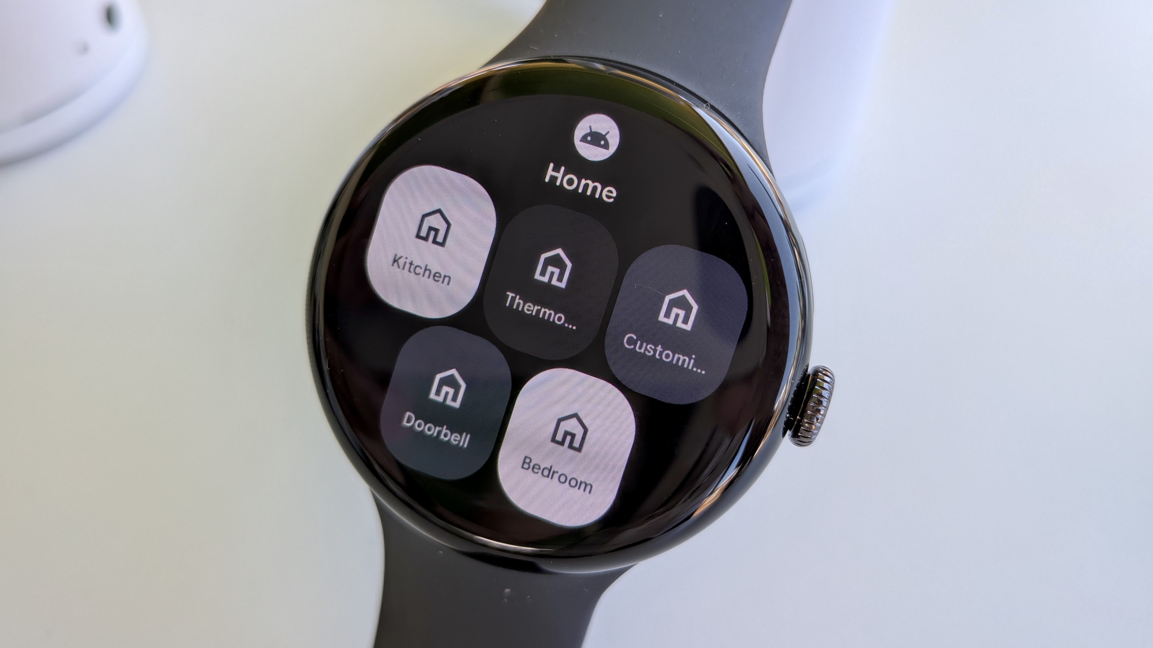 The new Google Home tile on Wear OS 6 on the Pixel Watch 3, showing options to check your Kitchen, Thermo, Custom, Doorbell, and Bedroom controls.