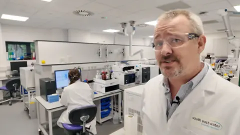 A man with grey hair and a beard wearing safety glasses and a white coat stands in a laboratory