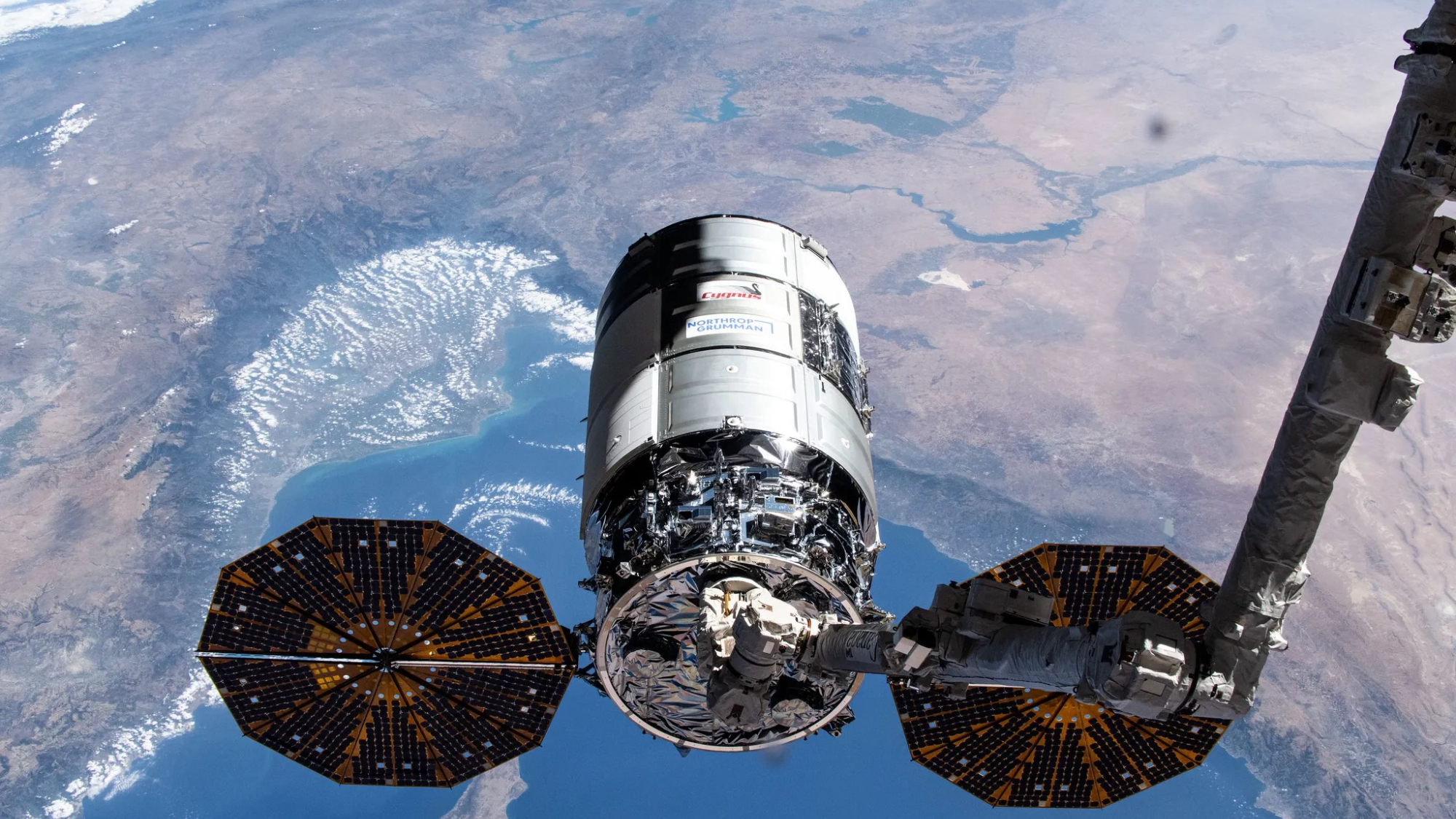 a cylindrical spacecraft with two octagonal solar arrays on the end of a multi-segmented arm, with earth visible far in the background