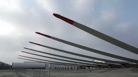 Seven large turbine blades are propped-up at a large open factory site. 
