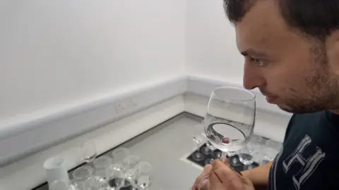 A man in a black top brings a wine glass containing a sample of water up to his nose to test the aroma of a sample of drinking water