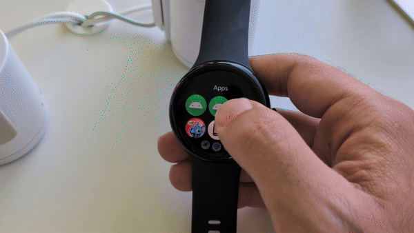 A Pixel Watch 3 held in hand showing the app drawer; the gif shows the holder scrolling up and down with his thumb, with apps shrinking at the top and bottom of the display.
