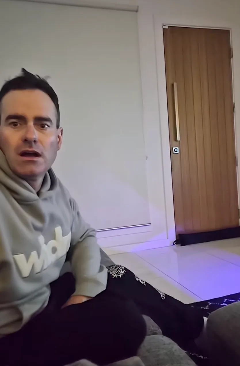 A person wearing a hoodie and patterned pants is sitting indoors, looking towards the camera with a surprised expression