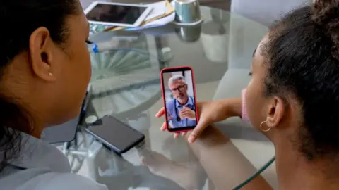 Getty Images Two women, seen from behind, hold a phone and speak to it. A doctor wearing a stethoscope round his neck is on the screen