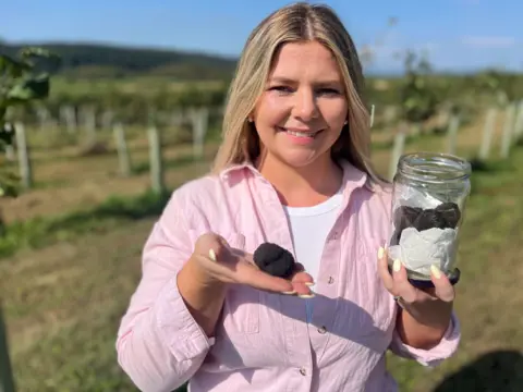A woman with blonde hair and white t shirt with a pink shirt is holding a single truffle in her right hand and a jar of them in her left.  She is standing in front of a junior orchard with blue skies.