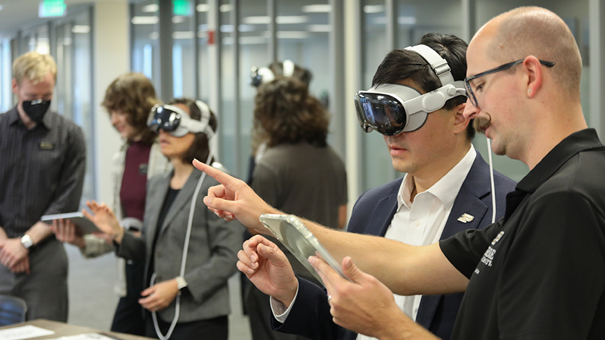 Purdue President Mung Chiang wears a virtual reality headset while another man reads from a tablet and points in front of Chiang; in the background, others wear headsets and look at tablets