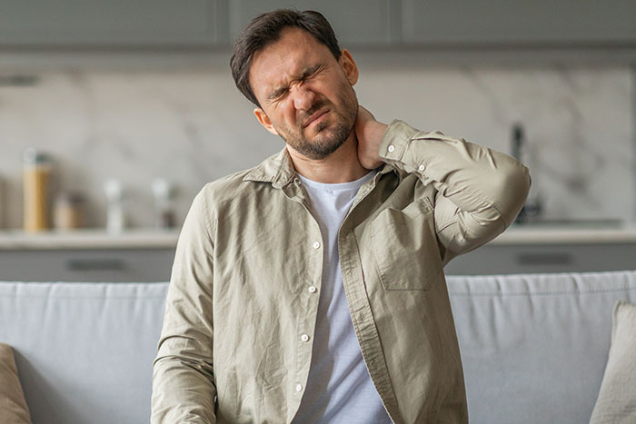 Man in casual shirt sitting on couch, holding neck in pain, illustrating struggles with chronic illness and relief attempts.
