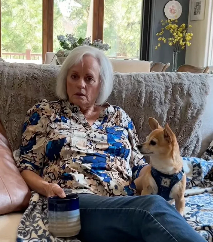 An elderly woman sits on a couch with a small dog beside her, holding a mug. She wears a patterned blouse