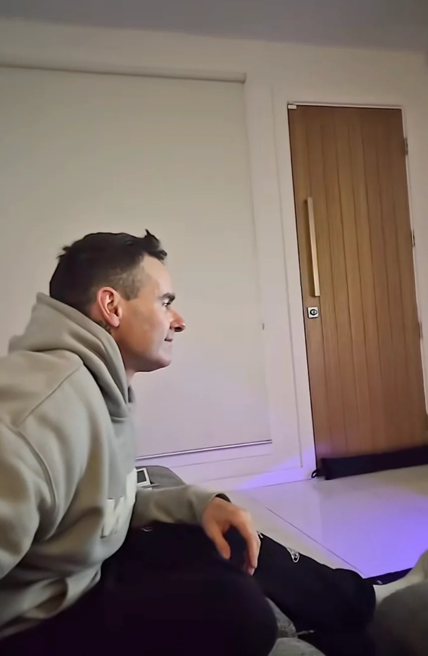 Person sitting on a couch wearing a hoodie, looking towards the right side. Interior of a room with a closed wooden door in the background