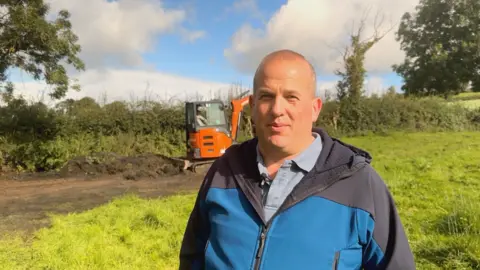 A bald man waring a blue shirt and blue and navy rainjacket, pictured from the chest up, stood in a field with green grass and hedges visible in background alongside a small orange digger. It is a cloudy day.