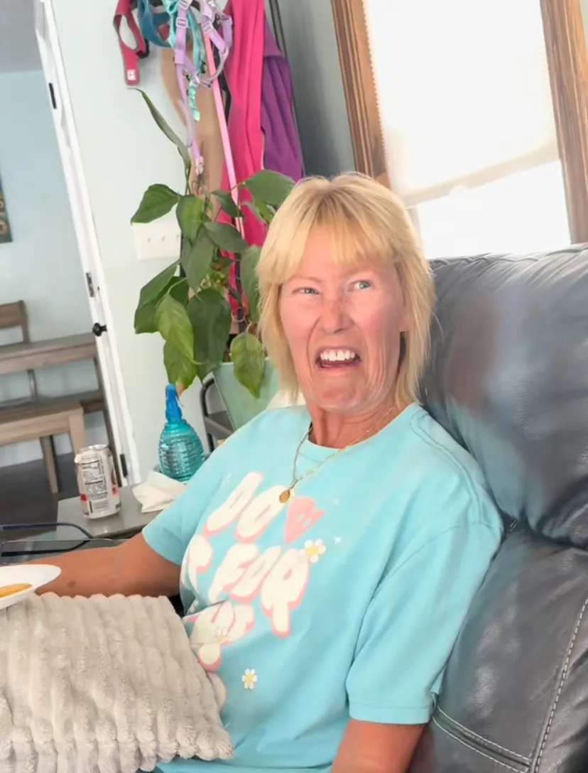 A person sitting on a couch makes a humorous facial expression, wearing a T-shirt with a playful design. A plant and household items are in the background