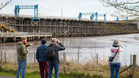 Damien Wootten Four people, two using binoculars, are pictured from behind, looking out from a path towards Dunston Staiths and the tidal basin. Dunston Staiths is a wooden structure similar in appearance to a pier. 