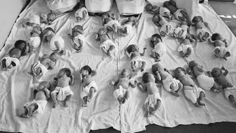 Getty Images A group of 30 babies all placed together on the floor of an NHS hospital in the 1960s. The babies are all on a large white sheet and facing in different directions, lying on the floor.