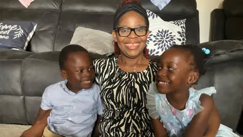 Perpetual with her son Osinachi and her daughter Sochika. They are sitting on the floor at home in front of a sofa. Perpetual has her arms around the twins. They are all smiling and looking happy.