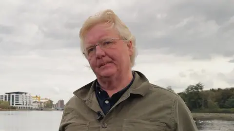 BBC A man with medium length blonde hair, wearing a khaki linen overshirt and a black undershirt, stood against a railing overlooking a body of water in Belfast. Trees and several structures, including the yellow harland and wolff cranes, are visible in the far distance.