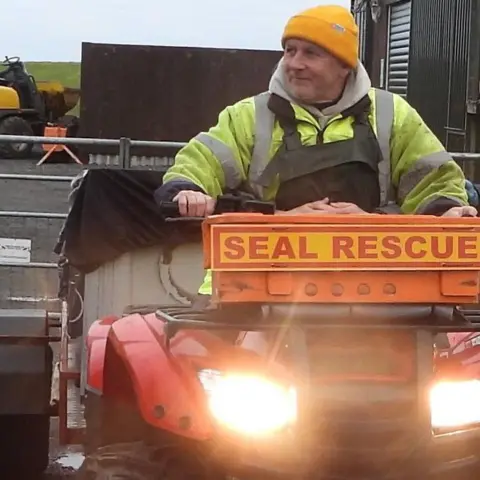 Caithness Seal Rehab and Release Centre Phil is riding a red quad bike. The machine's lights are on and it has an orange sign on the front with the words "seal rescue" in orange letters on a yellow background. The quad bike is pulling a small trailer with a sheet of black tarpaulin over the top of it. Phil is wearing green waterproof overalls over a yellow safety jacket. He is wearing a bright orange, woolly hat.