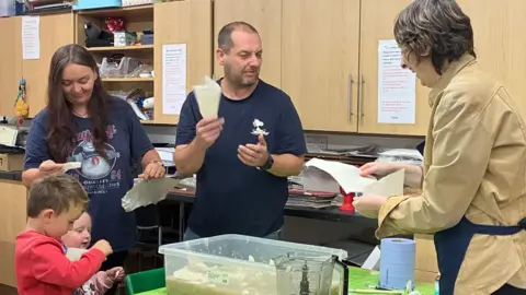 Natalie Brown demonstrates paper making to a family with two small children. They all hold scraps of paper and are putting them into a plastic tub filled with water