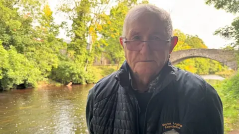 A man with short grey hair wearing glasses and a black jacket that says 'Moyola Angling Association'. He is stood beside a murky river with a stone footbridge and green trees in the background.