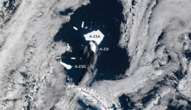 Angular white icebergs float atop dark blue ocean water. A long, white island appears near the bottom. Most of the scene is covered by clouds, except over the icebergs.