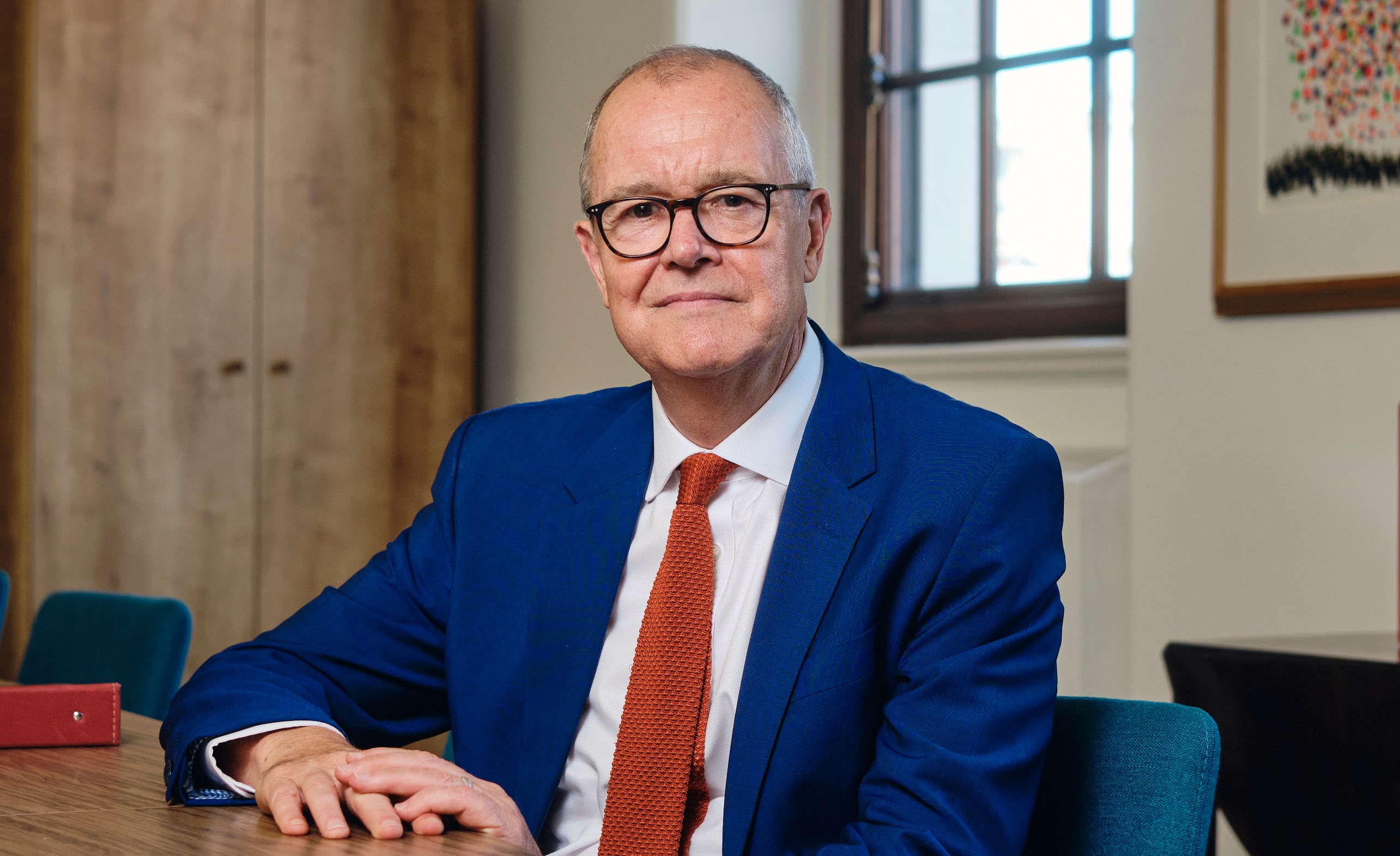 Lord Patrick Vallance seated at a wooden table in a blue suit and orange tie.