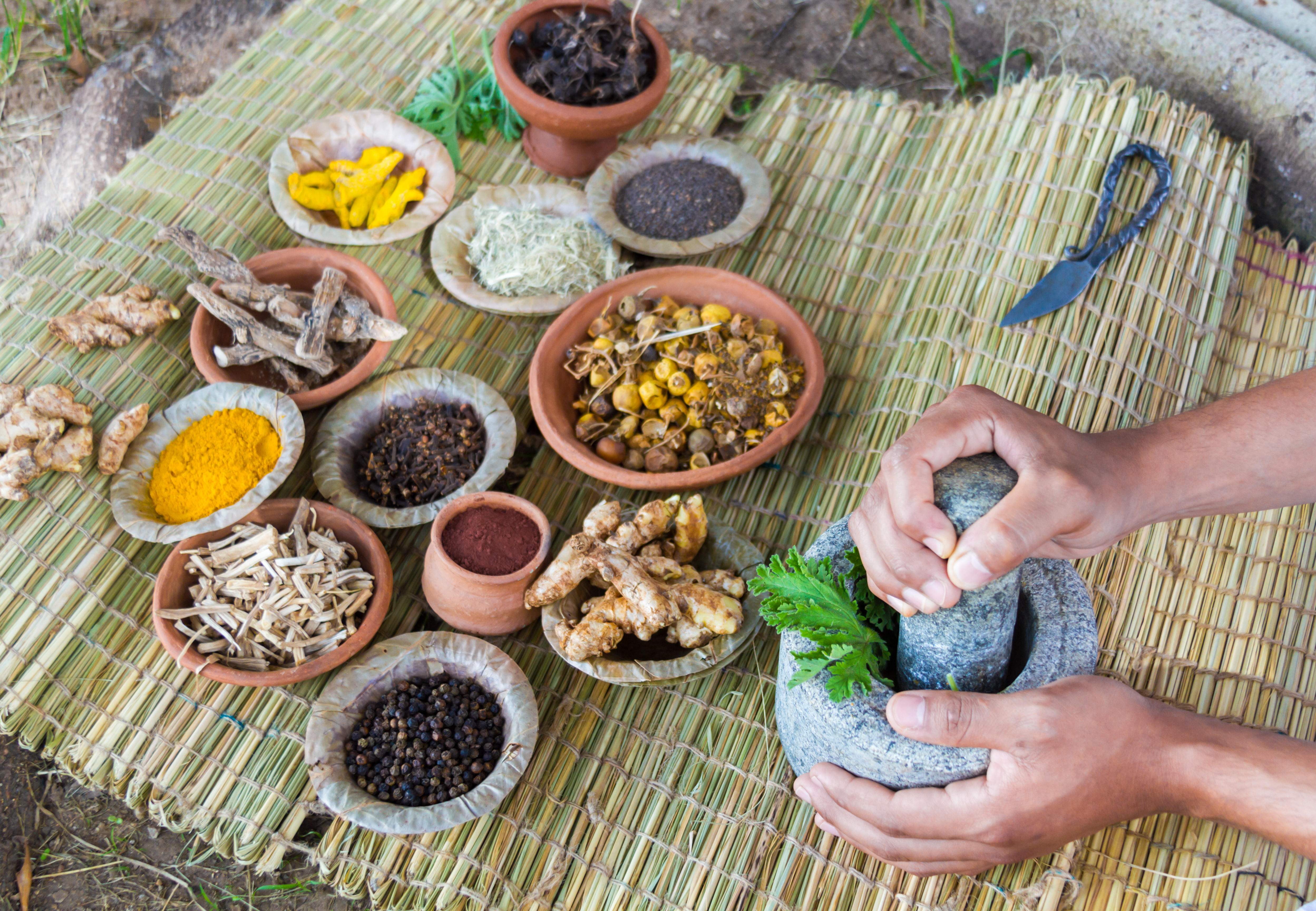 A person's hands using a mortar and pestle to grind green leaves next to bowls of various Ayurvedic herbs and spices on a woven mat.