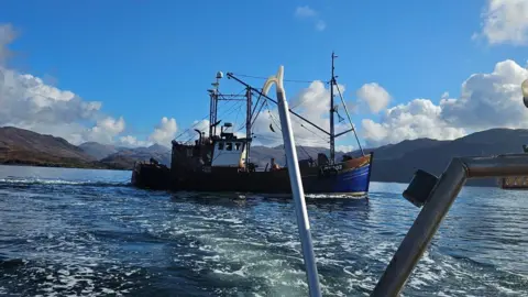 Bally Philp A blue boat with red trim is seen passing through the water with mountains behind and a blue sky